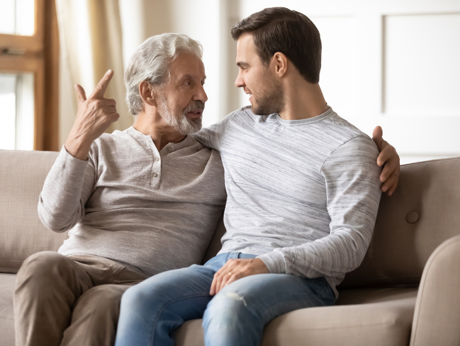 Two people sit side by side on a couch with their arms around each other's shoulders while engaged in conversation.