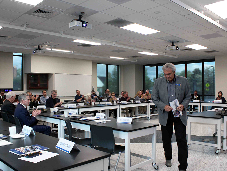 Man in a suit walks away from a podium while a group of 18 seated individuals applaud.