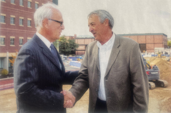 Two men in suits shake hands while standing in front of a construction site.