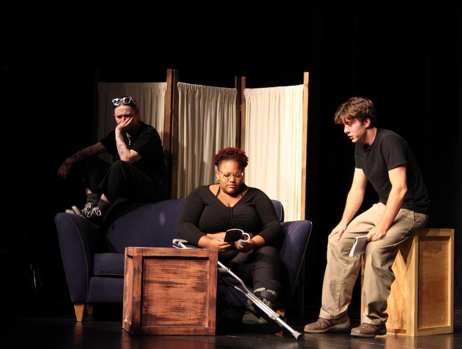 Three performers on stage - all have somber expressions.
