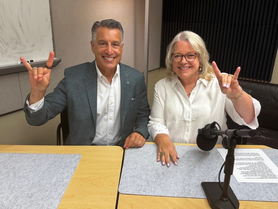 Tami Brancamp sits next to President Sandoval in the podcast recording studio, while making the Wolf Pack hand sign. 