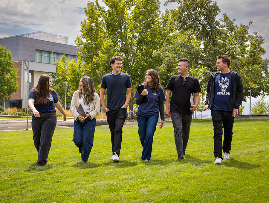 Six students walking and talking on a grassy field with laptops in hand.
