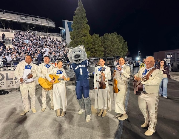 Performers and musicians dressed in traditional attire stand with Alphie the mascot on the field at Mackay Stadium during a football game.