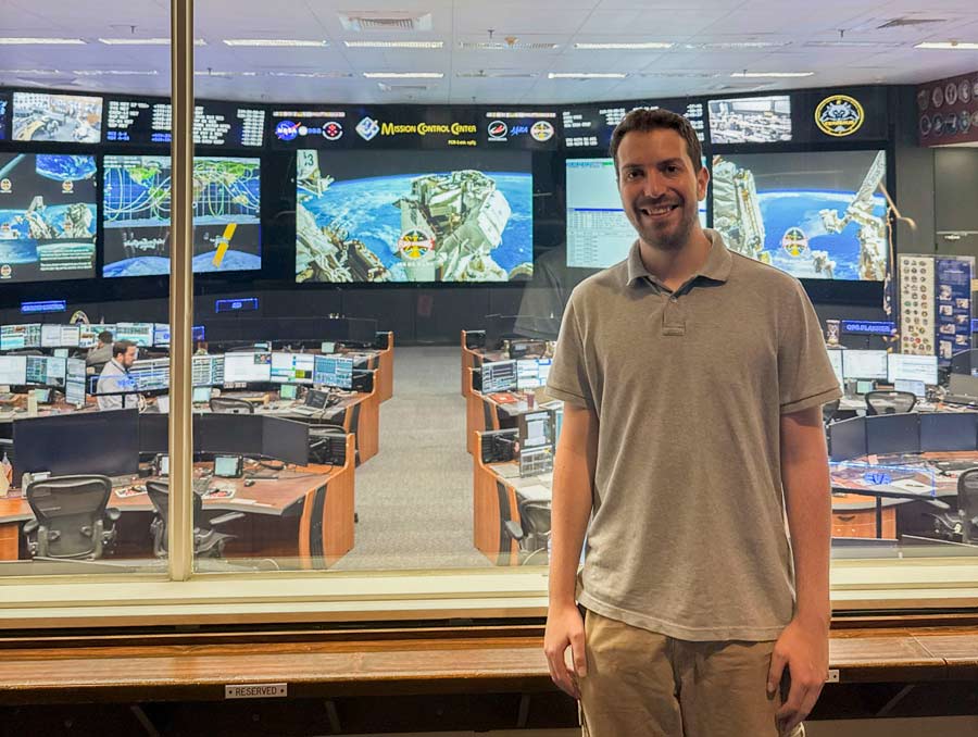 A man stands, smiling in front of a window looking into a room with desks and computers arranged toward large screens at the front of the room which say "Mission Control Center".