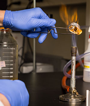 A close-up of a gloved hand lighting a bunson burner in a laboratory setting.