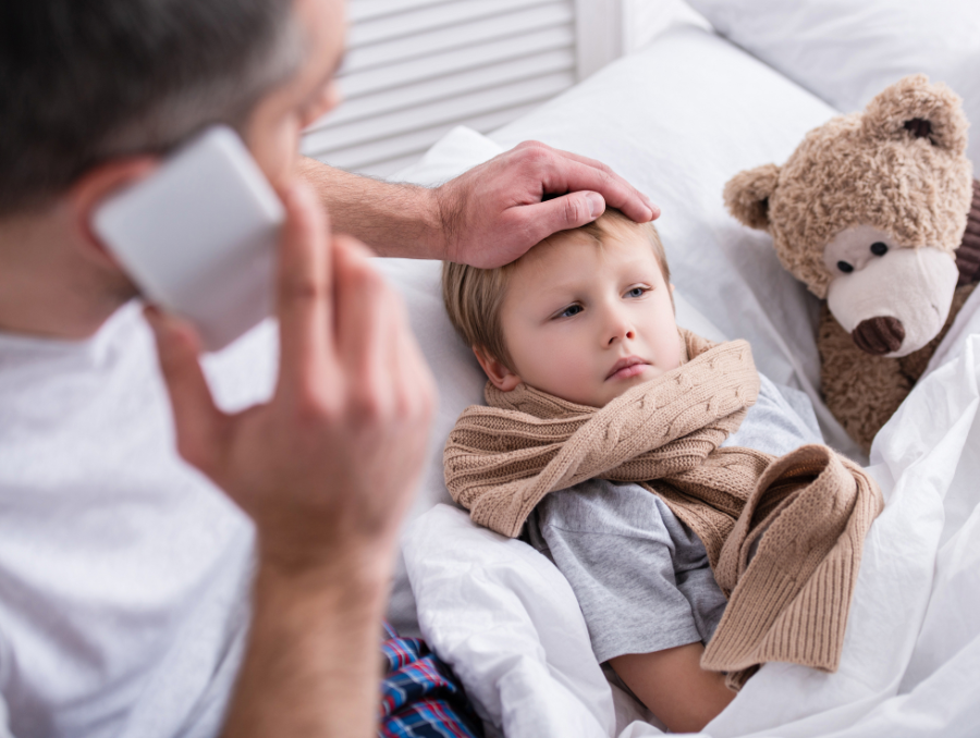 Sick child in bed with dad touching forehead and on the phone. There is a teddy bear next to the child.