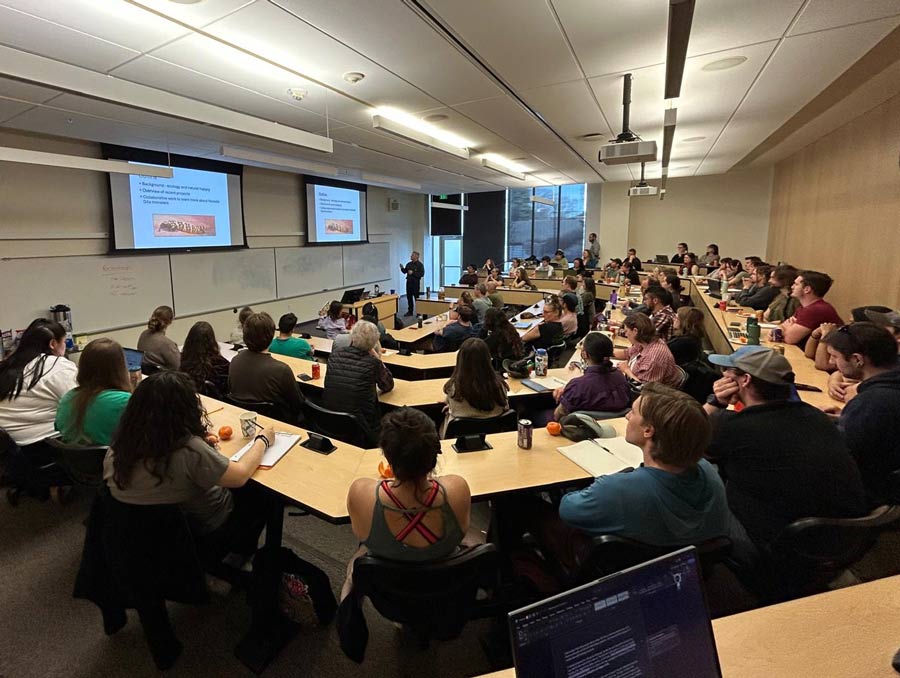 A packed classroom with a speaker at the front showing a presentation on two projector screens.