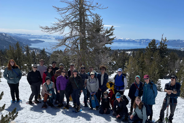 A group of people in winter attire and snowshoes pose for a photo on a snowy alpine hillside with Lake Tahoe in the background.