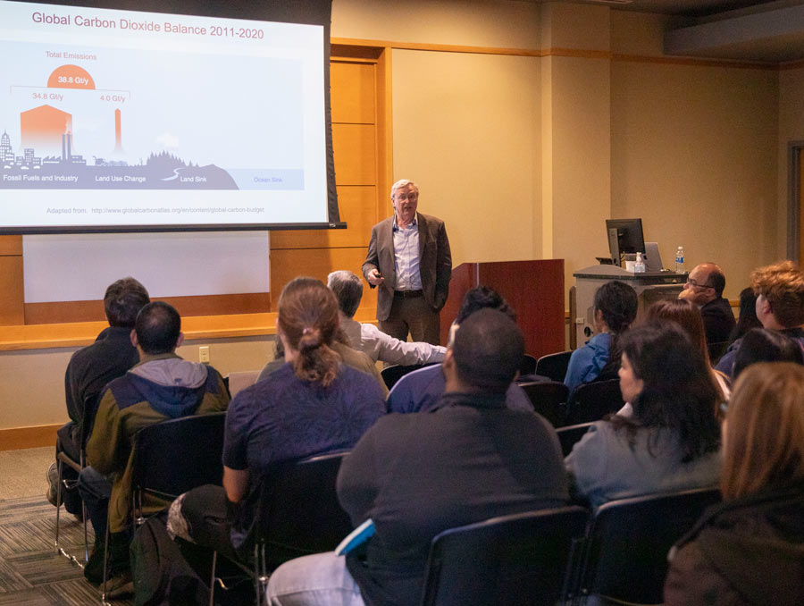 A man speaking in front of an audience with a presentation on a projector behind him.