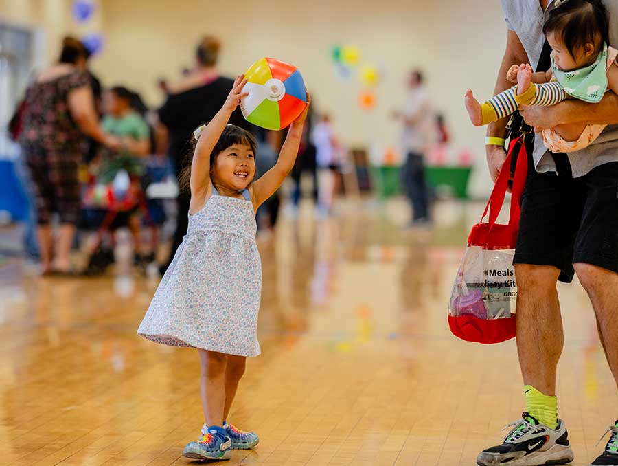 A smiling child holds a brightly colored inflatable beach ball above her head, reaching towards her parent and sibling.