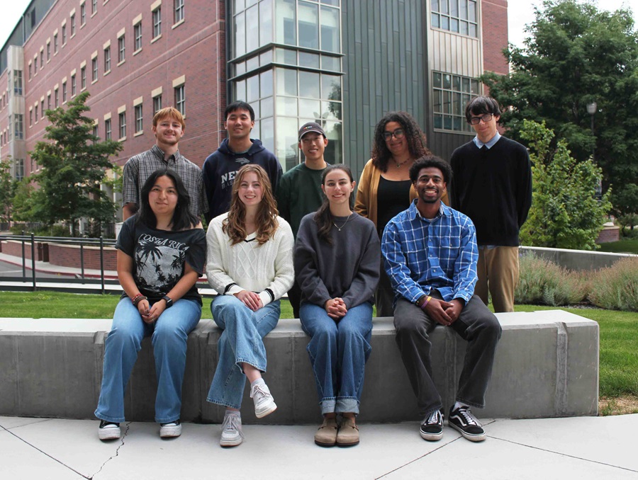 Nine students pose outdoors for a photo, four sitting on a concrete bench and five standing behind them.