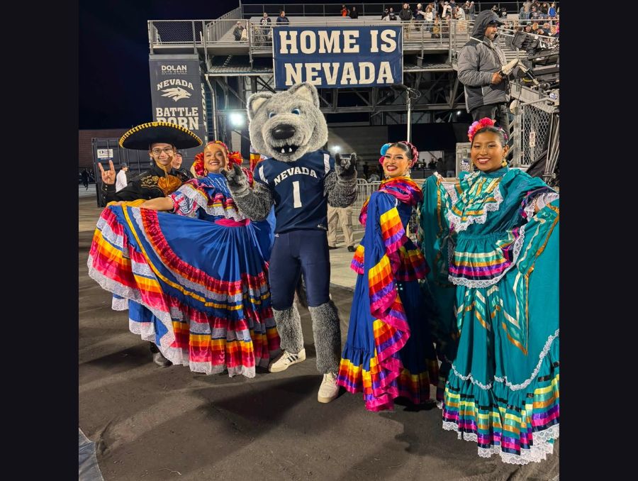 Folklorico dancers in colorful dresses posing on the field at Mackay Stadium with Alphie the mascot.
