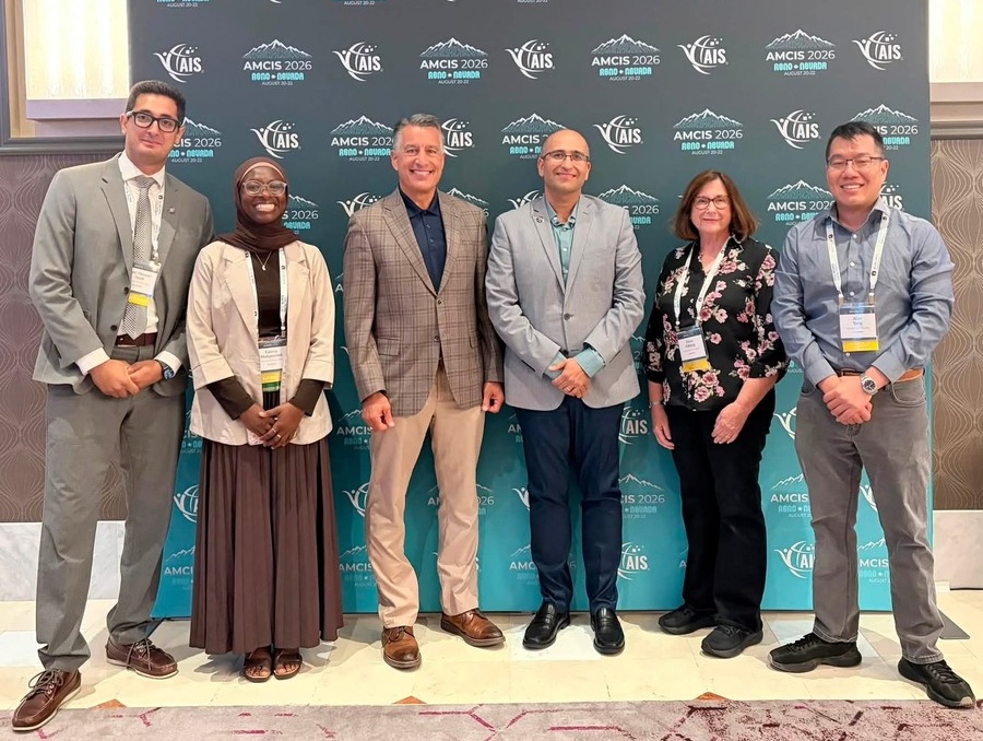 Six AMCIS 2026 attendees stand together in front of an AMCIS 2026 Reno conference backdrop. From left are Amir Yazdi, Fatima Mohammed, University President Brian Sandoval, Jason Talaei, Dana Edberg and Alan Yang, all wearing conference badges.