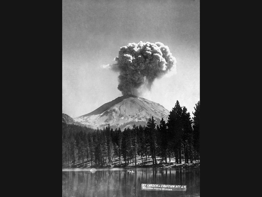 A black-and-white film photo of a volcanic eruption. The volcano, with a large plume of ash, is in the background of the photo, with an alpine lakeshore in the foreground. "No. 2" is handwritten on the photo in white ink, along with "Mt. Lassen in Eruption Oct. 6 '15" along with an illegible copyright note.