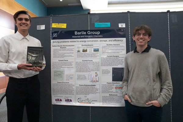 Two men stand in front of an academic poster, smiling for a photo. One holds up a metallic rectangle.
