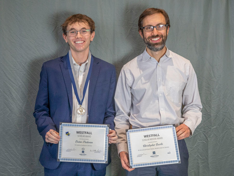 Two men smile for a portrait in front of a gray background. They are both holding certificates in front of them, and one of them is wearing a medal.
