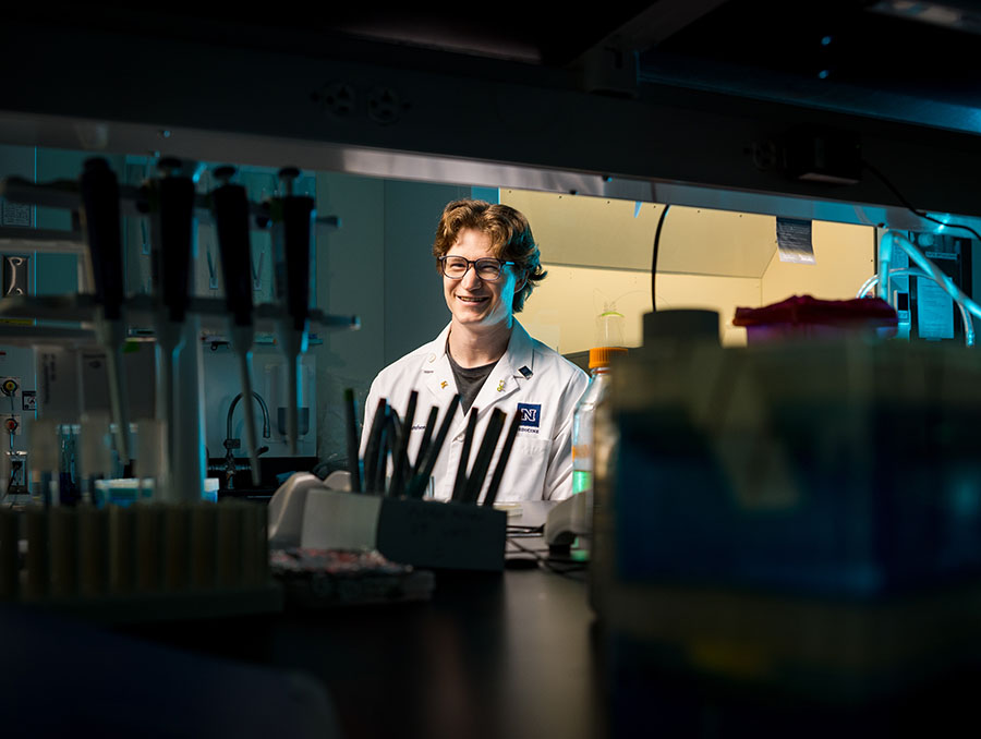 Ryan Gustafson in the laboratory looking through shelving with equipment such as pipets in the foreground.