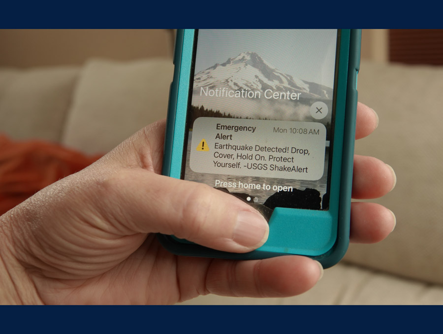 A hand holding a phone. A notification on the screen with a yellow alert symbol reads, "Emergency Alert. Earthquake Detected! Drop, Cover, Hold On. Protect Yourself. - USGS ShakeAlert"