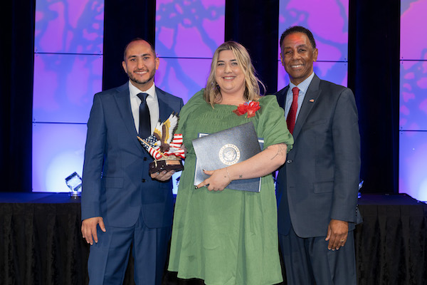 Sadie stands in a green dress on a stage between two men who are presenting her with awards.