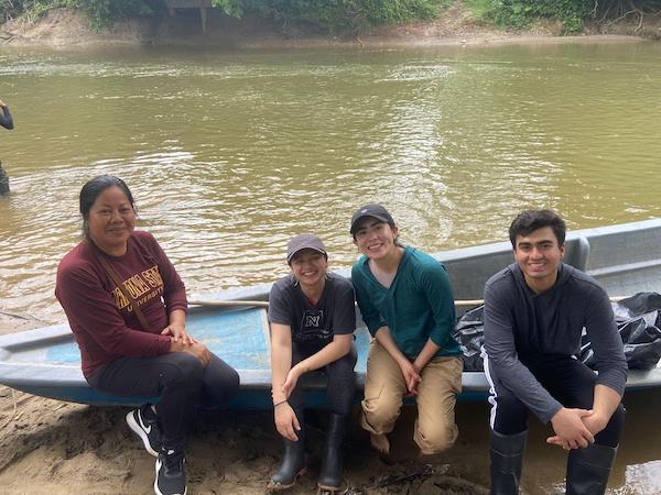 A group of four smiling young people sit in a boat on the edge of a green water river.