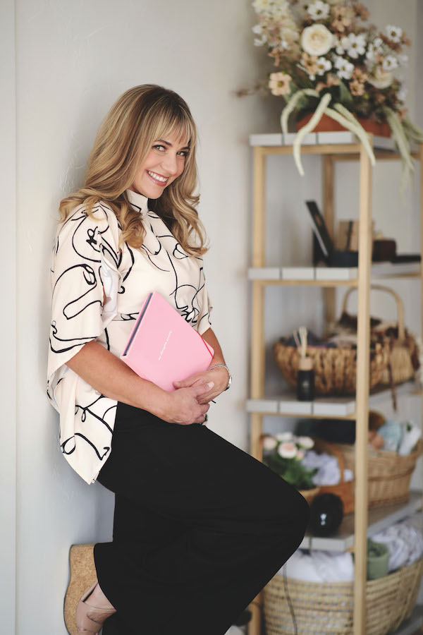 Joslyn leans against a wall carrying a pink notebook and smiles at the camera