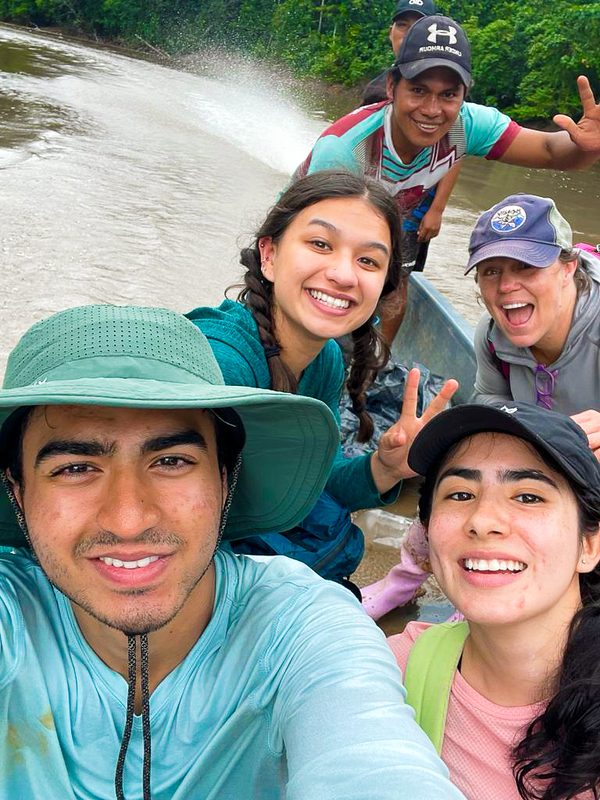 Six people seen smiling on a boat that is gliding down a river. Everyone is smiling and some are holding up peace signs.