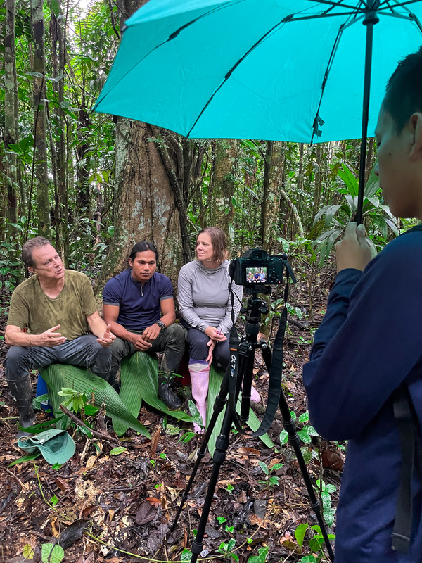 Three people sit on leaves in the jungle talking while in front of a camera on a tripod. You can see someone in the foreground holding an umbrella and watching the camera.