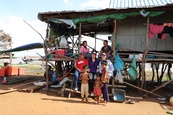 A family including four adults and three children pose with Lucy on the steps outside their fishing hut near the water.