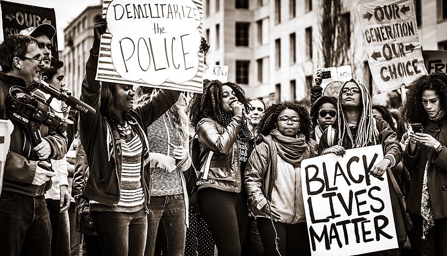 A group of people stand holding protest signs that read "Demilitarize the police" "Black Lives Matter" and "Our Generation Our Choice." The image is in black and white.