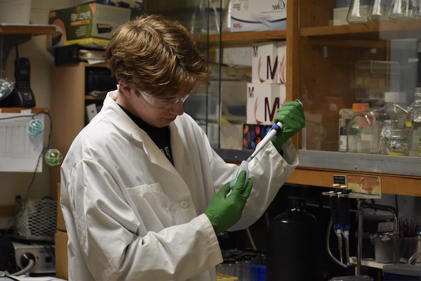 Jaeden in a lab holding a vial and using a syringe to add drops into it.