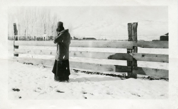 Snowy landscape with a wooden fence and a woman standing in winter clothing, facing away from the camera. Black and white.
