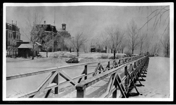 A historical photo in black and white of a snowy wooden bridge on Manzanita Lake with young, barren trees and University buildings in the background.