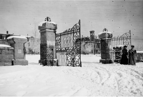A snowy landscape with iron gates and two women in clothing from the early 1900s with Morrill Hall in the background.