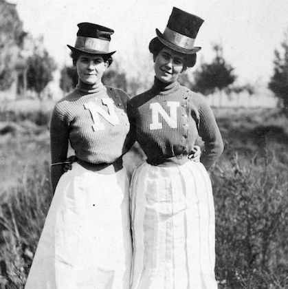 Black and white photo from 1904 of two ladies on campus wearing top hats, Nevada sweaters, belts, and long white skirts.