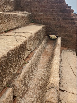 A stone staircase from close up where you can see that one of the steps is curved inward and made to drain out a small hole in the side walling of the stairs.