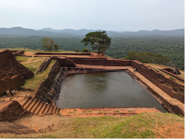 An overlook of a large manmade pond carved into a stone hillop with some grass and trees around the brown stone with a short set of stairs leading to it.