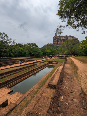 A large reflection pool carved into stone in the foreground with a looming stone, hilltop temple in the background.