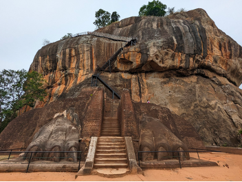 A large rocky hill topped with trees and a stone stair case in the middle with the two areas next to the staircase carved to look like lion's paws.