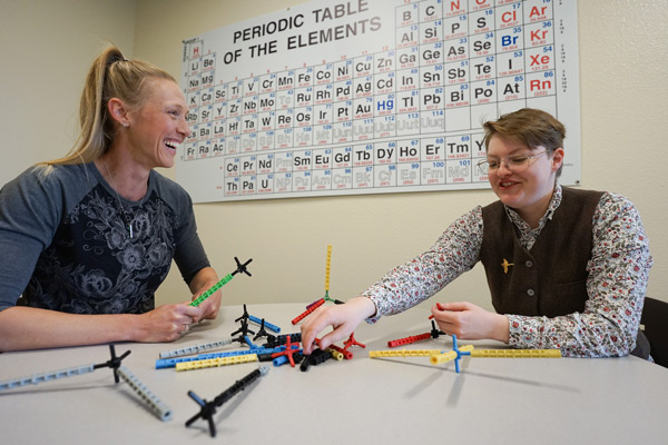 Lyndsay Munro sits with student Beck Neal as they use colorful materials to build molecular models, smiling.