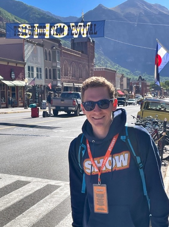 John wears a name badge and smiles broadly while standing on a historic street in Telluride.