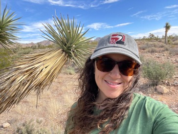 Ranae Sullivan standing outside in the desert with palm trees behind her while she smiles and wears a baseball cap and sunglasses.