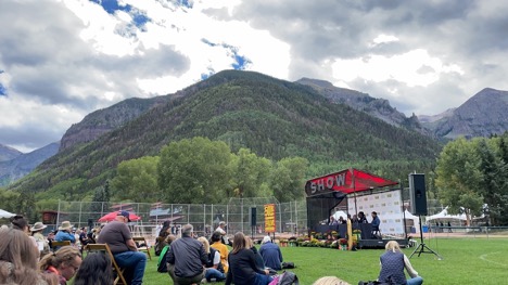 A group of people sit outside on green grass facing a stage.