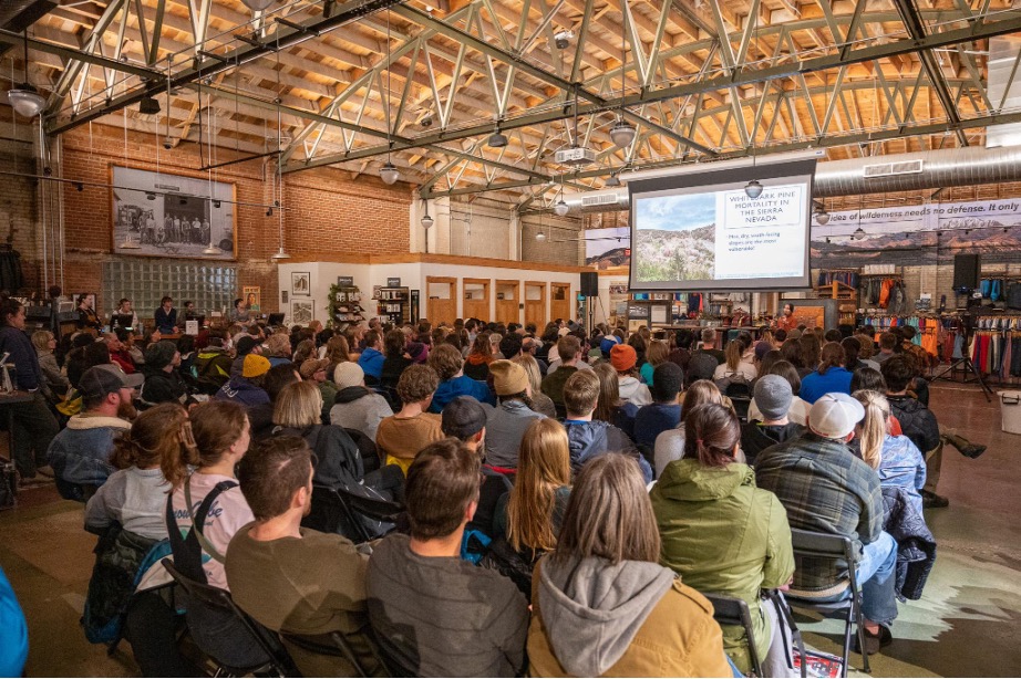 A very large crowd of people sitting in a warehouse listening intently to someone speaking with a presentation showing