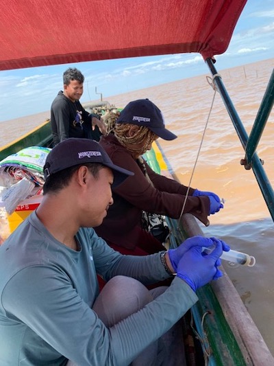 Two researchers and a boat captain on a brown river wearing gloves and collecting water over the edge of the boat.