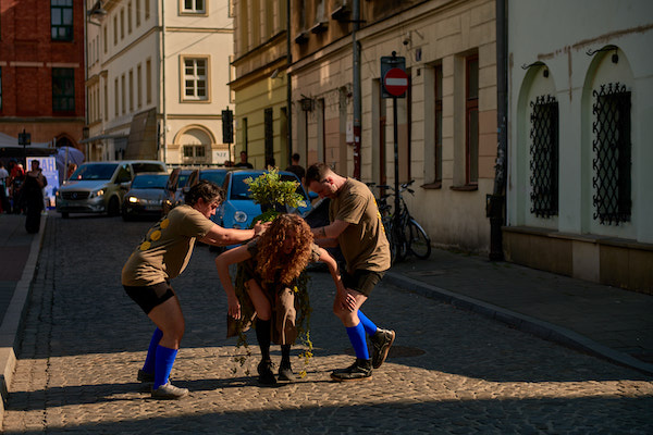 Three dancers in the middle of the street during a live performance. A woman is in the center, bending at the hips while a plant is placed on her back by two males on either side of her.