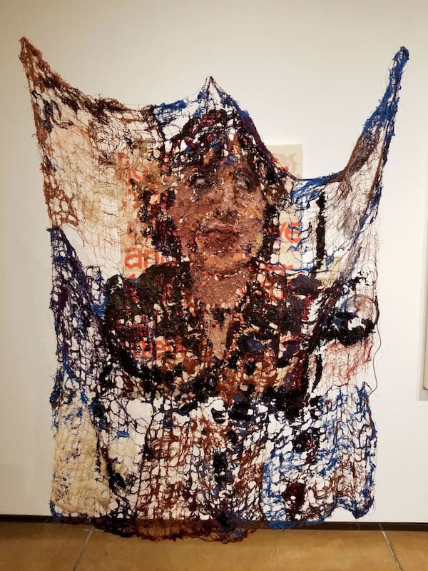 A piece of artwork hanging in a gallery. It looks like fabric with holes in it with the image of a woman's head and shoulder.