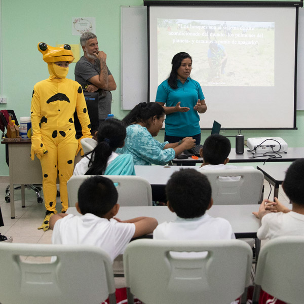 Three people stand in front of a classroom, including one dressed up as a yellow frog with black spots. One person is speaking in front of a projector screen.