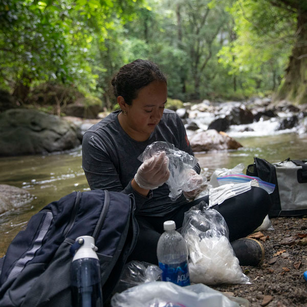 A woman sitting on a riverbank is wearing plastic gloves and holding a plastic bag in her hand.