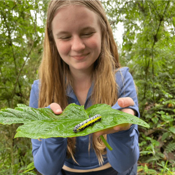A girl smiles and holds a large leaf with a large caterpillar on it.