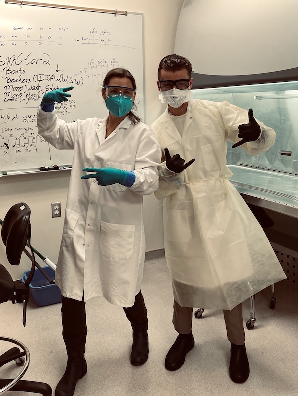 Berner and Gillis in a lab wearing protective lab equipment and making peace and hang loose signs with their hands.